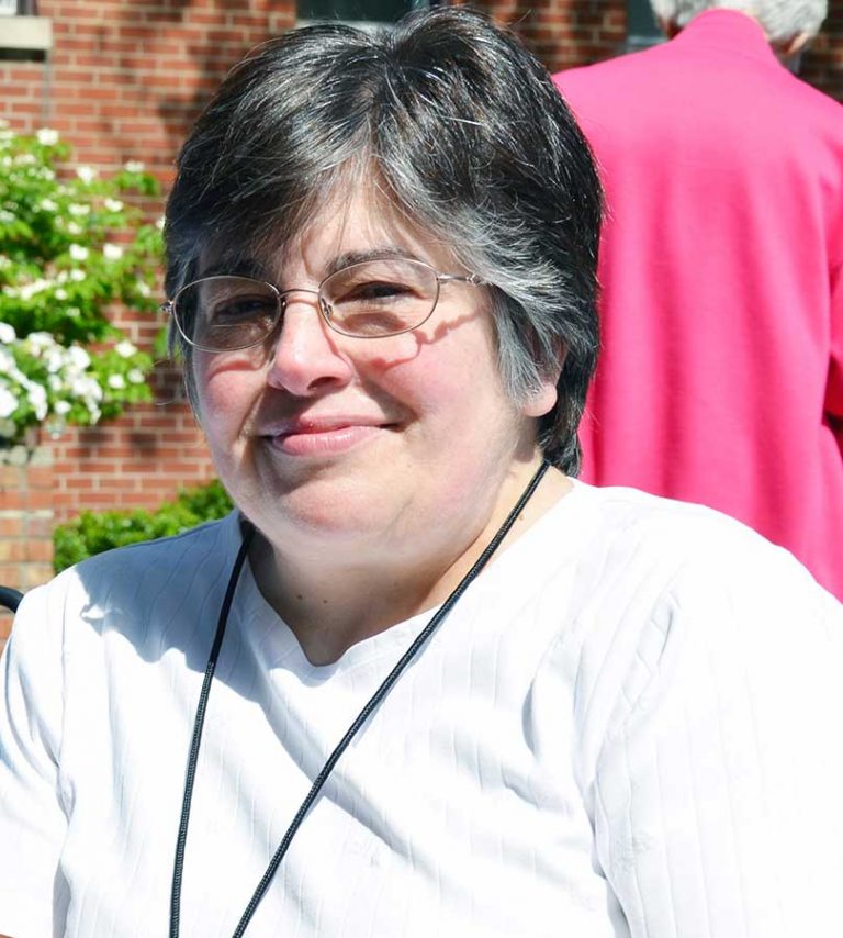 A woman, Mary Gutzwiller, with short dark grey hair, and wire-rimmed glasses wearing a white t-shirt and black lanyard smiling for a picture outdoors on a sunny day in front of a brick building with a well-manicured lawn and bushes.