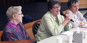 Three women pictured candidly sitting at a long table together, the woman in the middle is speaking into a black microphone to a crowd out of frame.