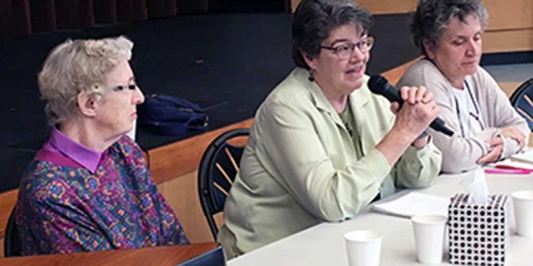 Three women pictured candidly sitting at a long table together, the woman in the middle is speaking into a black microphone to a crowd out of frame.