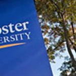 A royal blue with white lettering sign for Webster University with a background of large leafy trees.