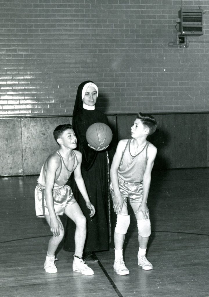 Two boys in basketball uniforms prepare to jump for a ball when the Sister tosses it