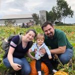 A young family: a man, woman, and young child smiling together for a group picture while crouching in a pumpkin patch outdoors.