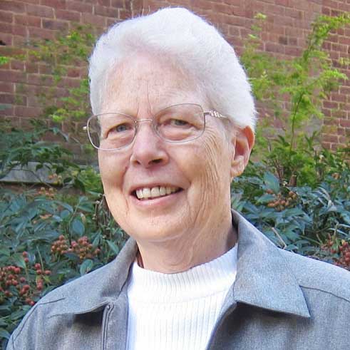 A woman with short, white hair and wire glasses smiling brightly for a headshot picture wearing a white t-shirt and a grey collared jacket outside in front of a brick building and greenery.