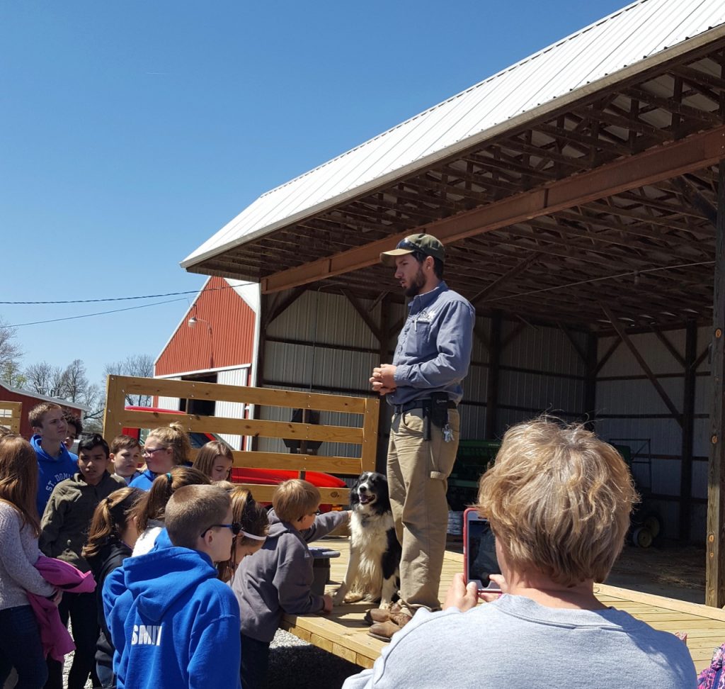 Students gather and listen to man standing on a hayrack.