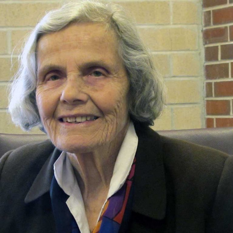A woman with shoulder-length grey hair wearing a patterned blouse with a white collar and a black blazer smiling for a headshot picture while sitting on a black leather seat in front of a brick wall.