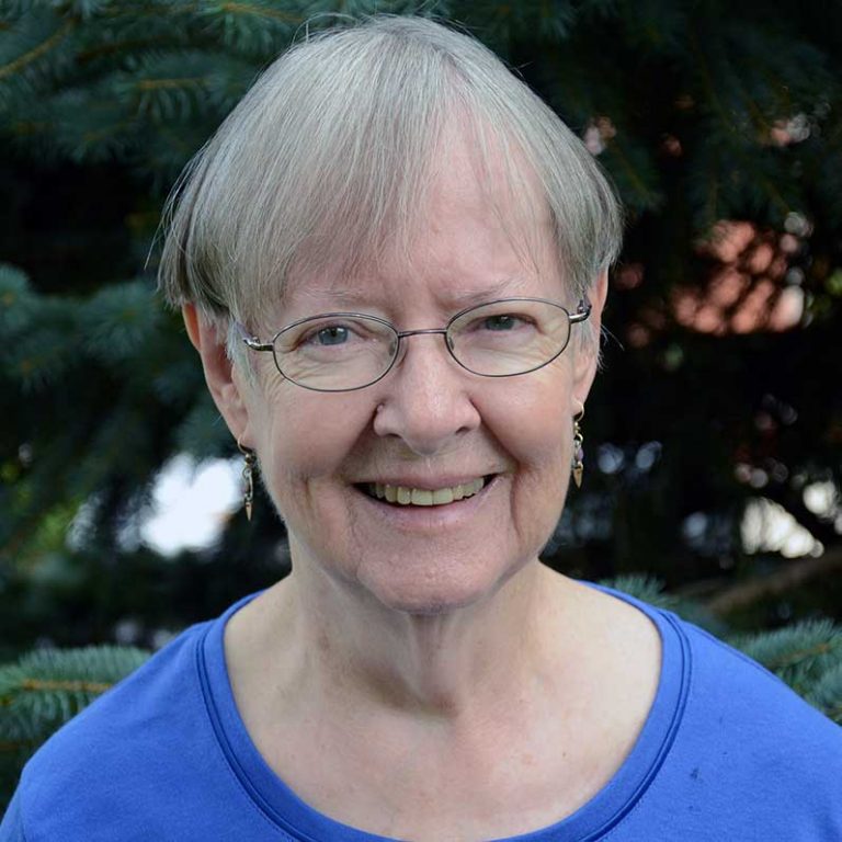 A woman with short grey hair and round wire glasses smiling brightly for a headshot picture outdoors in front of spruce tree greenery wearing a bright blue blouse.