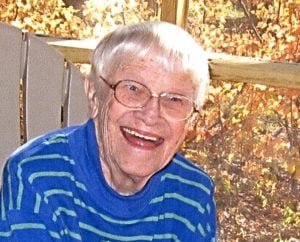A woman with short, grey hair, smiling for a picture while sitting in a lawn chair outdoors in autumn. She is wearing a dark blue striped sweater.