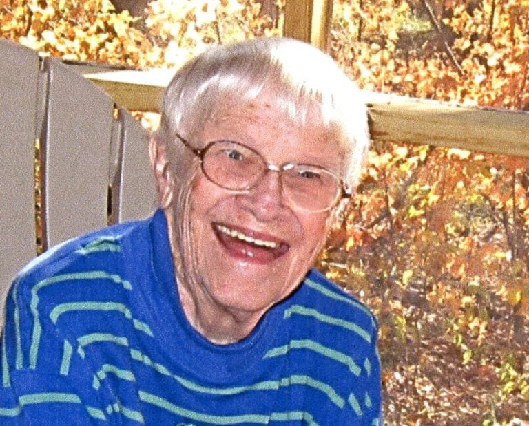 A woman with short, grey hair, smiling for a picture while sitting in a lawn chair outdoors in autumn. She is wearing a dark blue striped sweater.