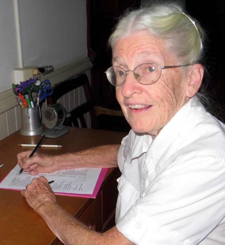 A woman with white hair and oval glasses smiling while sitting at an oak wood desk and writing a letter on lined paper on top of a bright pink folder. She is wearing a short-sleeved, white, collared, button up shirt and patterned bottoms and the oak desk she is sitting at is indoors.