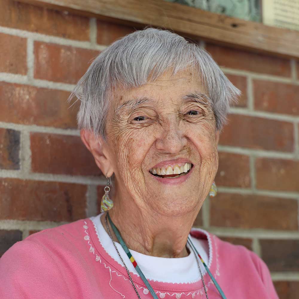 A woman with short grey hair wearing a white undershirt, pink sweater, beaded necklace, and dangly earrings smiling brightly for a headshot picture while leaning against a red brick wall.
