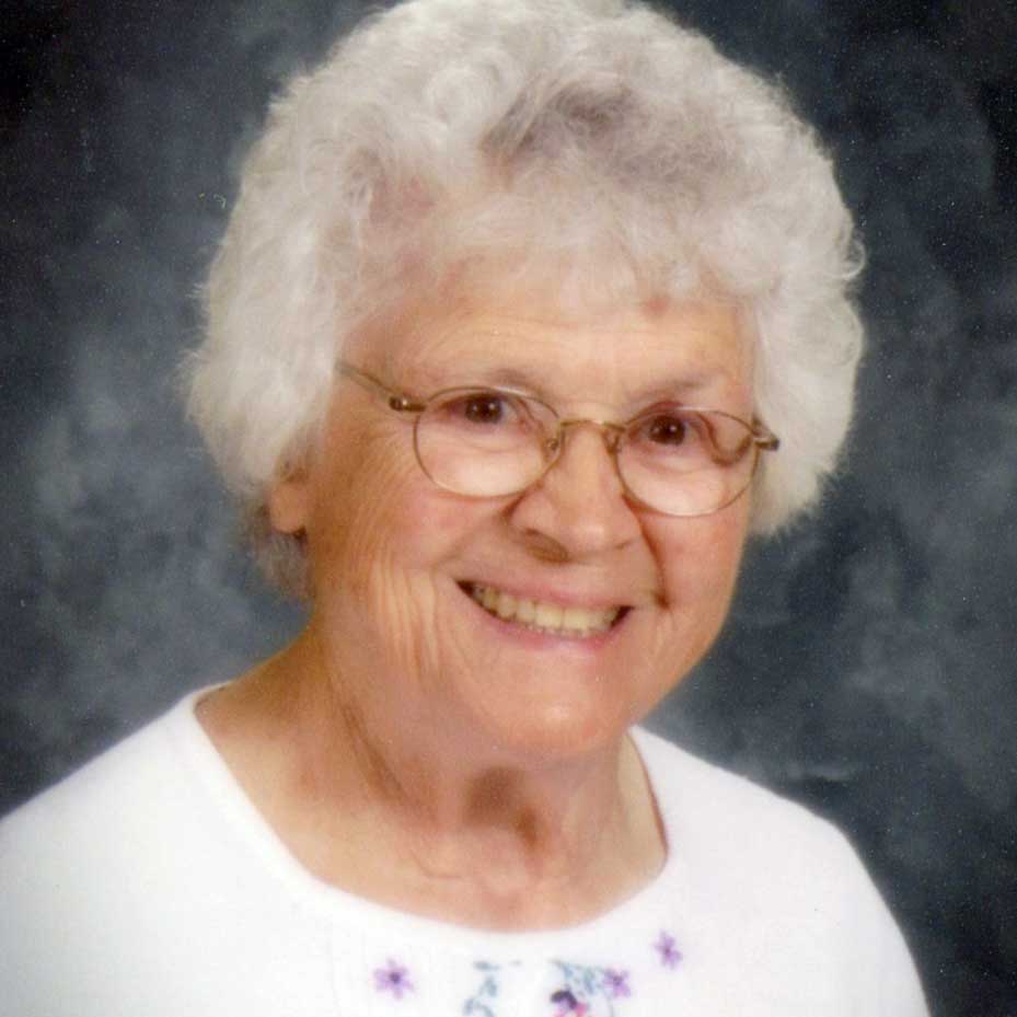 A woman with short white hair and oval wire glasses wearing a white blouse with dainty floral patterns smiling brightly for a portrait headshot picture with a black and grey vignette background.