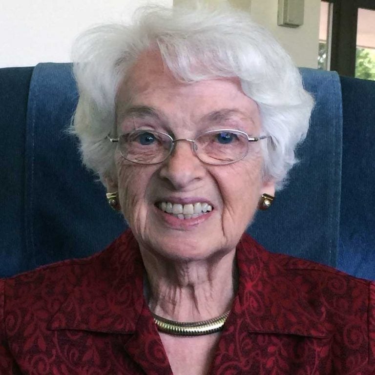 A woman with short white hair and round wire glasses smiling brightly for a headshot picture while wearing a red collared blouse and a thick gold necklace while sitting in a velvet blue recliner chair in a living room.