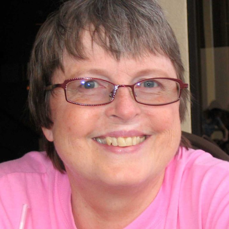 A woman with short dark grey hair and oval dark-framed glasses smiling brightly for a headshot picture while wearing a flamingo pink shirt.