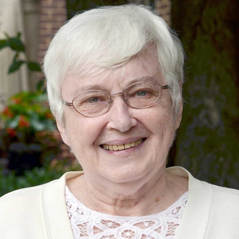 A woman with short white hair, oval shaped glasses, and blue eyes wearing a white blouse with lace detailing smiling for a headshot picture outside of a brick house with greenery behind her.