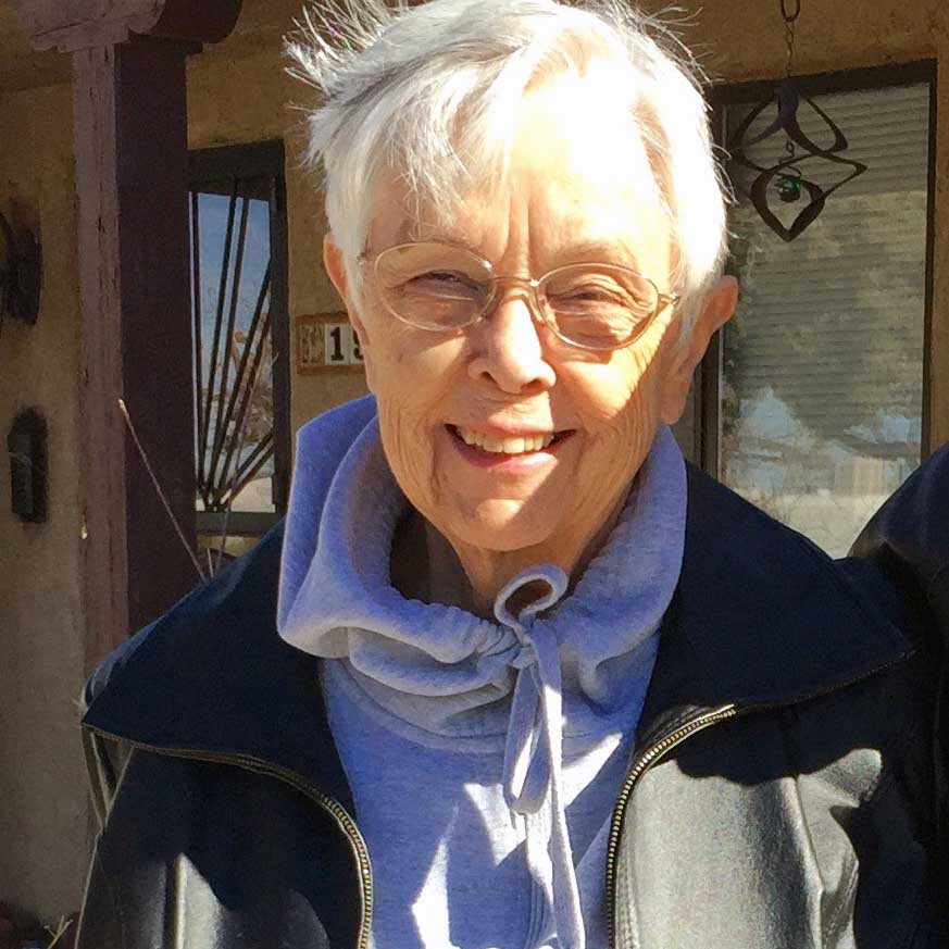 A woman with short grey hair and round glasses smiling for a headshot picture outdoors on a sunny day in front of a house. She is wearing a light purple sweatshirt and a black jacket on top.