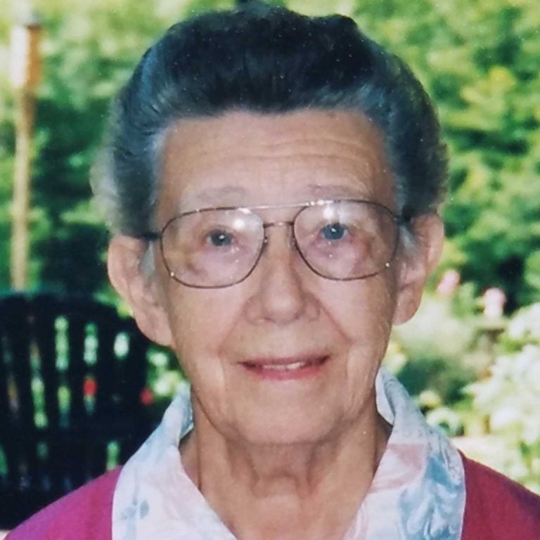 A woman with dark grey hair pulled back and wire glasses wearing a pink sweater and a white floral patterned blouse smiling softly for a headshot picture outdoors on a sunny day in front of green trees.