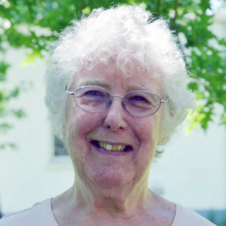 A woman with short white hair and round wire glasses wearing a white blouse smiling brightly for a headshot picture outdoors on a sunny day underneath a green tree.