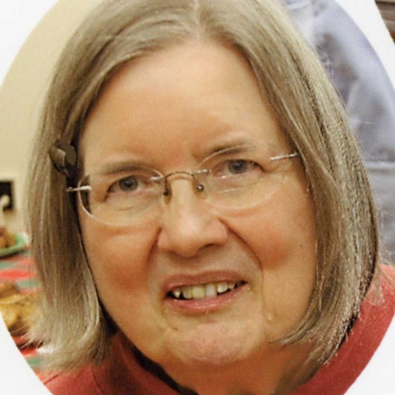 A woman with shoulder-length light brown hair and glasses wearing a terracotta sweater smiling softly for a headshot picture. The picture has an edited white oval frame framing her face.