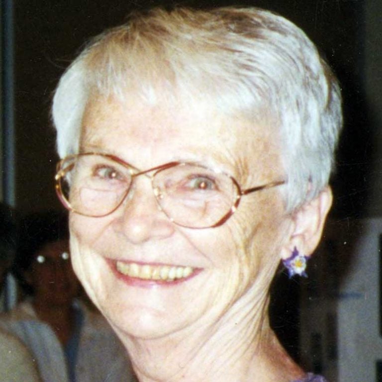 A woman with short white hair, dangly earrings, and glasses smiling brightly for a flash headshot picture in a dimly lit room.