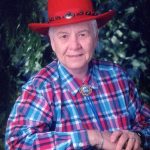 A woman with short white hair wearing a blue and red plaid collared shirt and a red cowgirl hat with silver detailing smiling for a portrait picture in front of a dark greenery background.