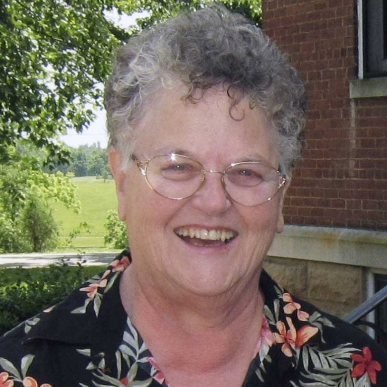 A woman with short grey hair and round glasses wearing a black floral patterned blouse smiling brightly for a headshot picture outdoors on a sunny day in front of a red brick building under a green tree.