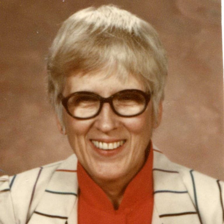 A woman with short grey hair and oval black-framed glasses wearing a red blouse and a white blazer with thin black stripes smiling brightly for a portrait headshot picture in front of a plain beige background.