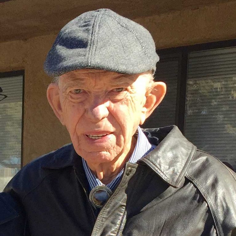 A man wearing a dark grey flat hat, a light blue collared shirt, and a worn leather jacket smiling brightly outdoors on a sunny day.
