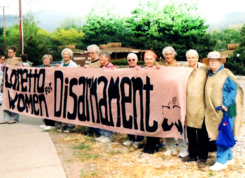 Loretto Community members wearing sackcloth stand by the sidewalk with a banner that reads "Loretto Women for Disarmament."