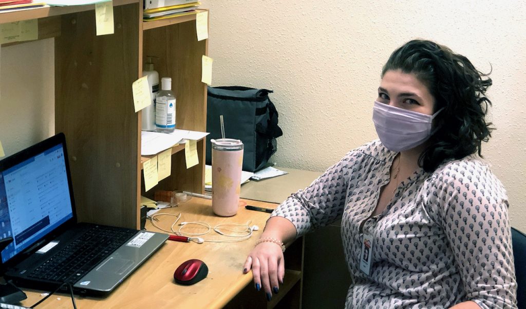 A young woman in a mask sits at her desk in front of a laptop.
