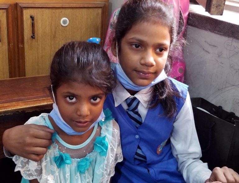 Two young Pakistani girls, one in a school uniform and one in a lacey dress, sit together.