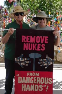 Two women, wearing hats and sunglasses and holding a banner that reads 'Remove nukes from "Dangerous Hands,"' stand in front of thousands of hanging paper cranes.
