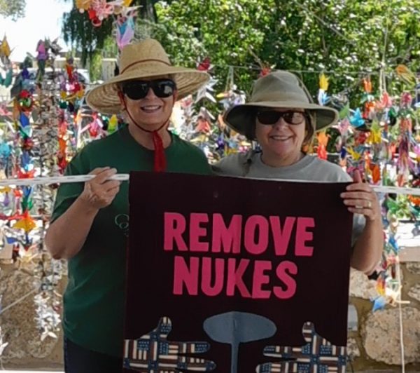 Two women in hats and sunglassed hold a "Remove Nukes" banner in front of strands of paper cranes.