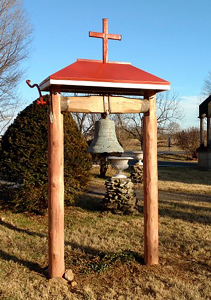 A large bell hangs outside on a wooden beam held up by two wooden posts. A cross sits atop the beam.