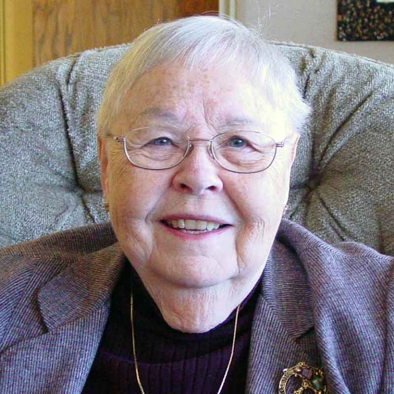 A woman with short white hair and oval wire-framed glasses wearing a black blouse, gold chain necklace, and a purple blazer smiling brightly for a headshot picture while sitting in a grey recliner chair in a living room.