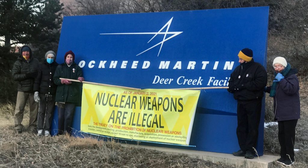 Five people in winter clothing and face masks stand next to the Lockheed Martin sign holding a banner with the text "Nuclear Weapons are Illegal"