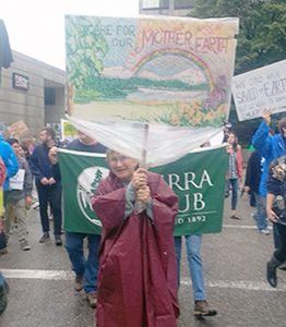 Woman in rainponcho marches with poster reading "Care for Our Mother Earth."