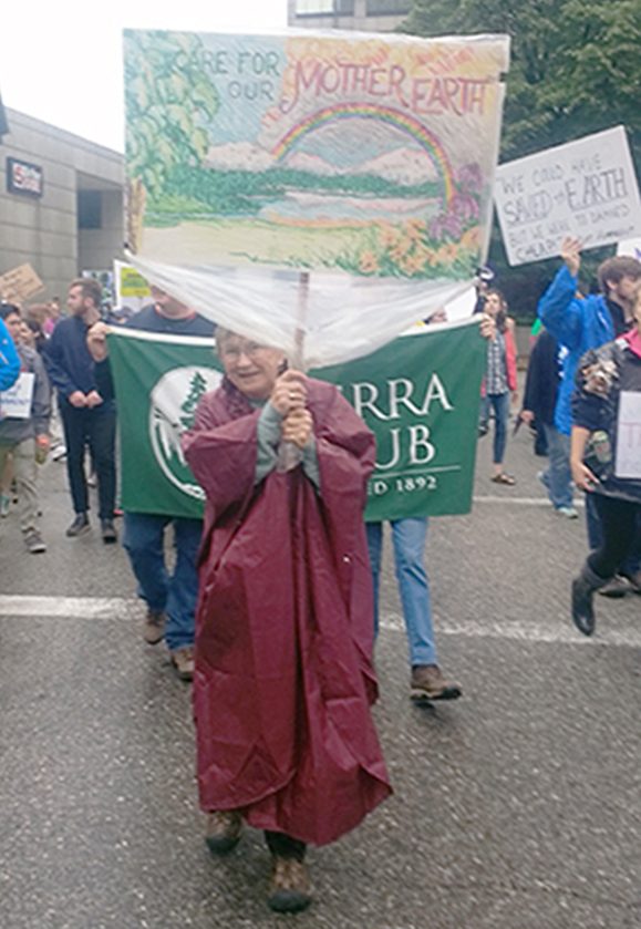 Woman in rain poncho marches with a sign saying "Care for our Mother Earth."