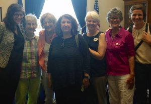 Seven women stand in front a window with a US flag behind them