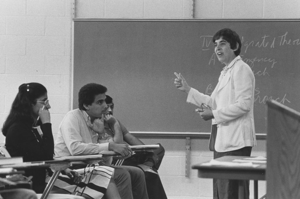 A woman stands at the front of a blackboard. Three students sit in desks, listening to her lecture.