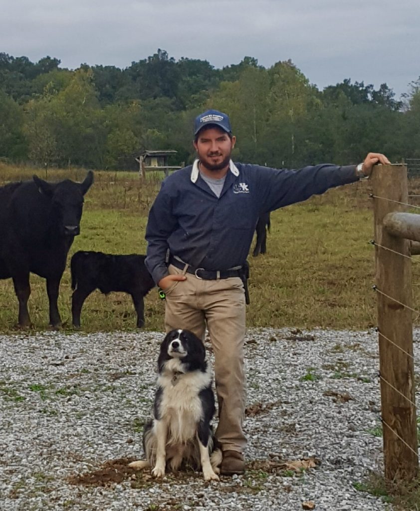 A man stands, with an arm out on a fence post, with a black and white working dog sitting in front of him.