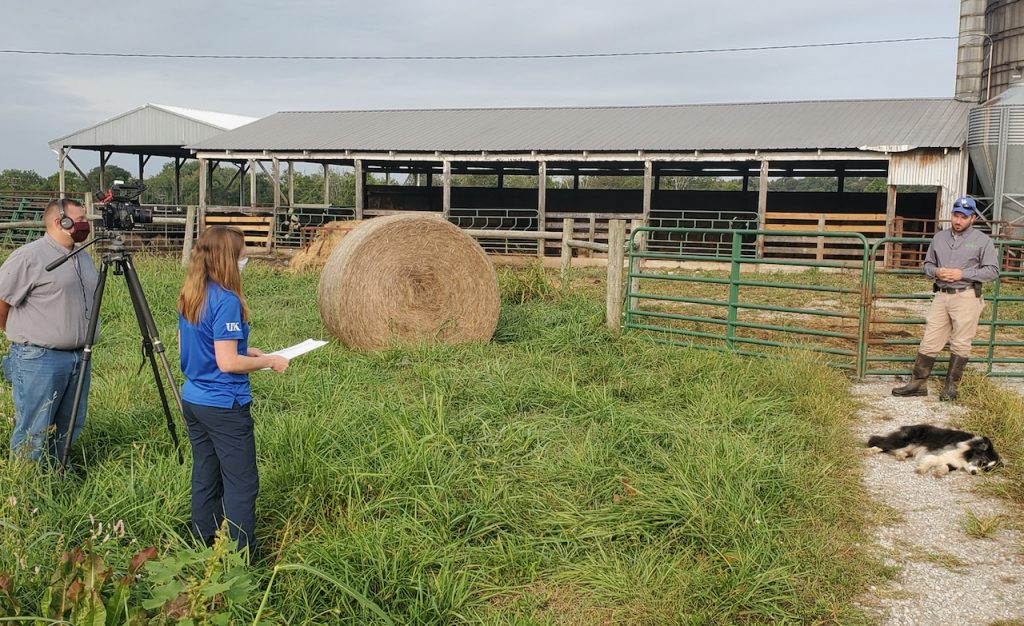 Masked cameraman and interviewer film the farmer from a COVID-safe distance in front of a livestock paddock and barn.