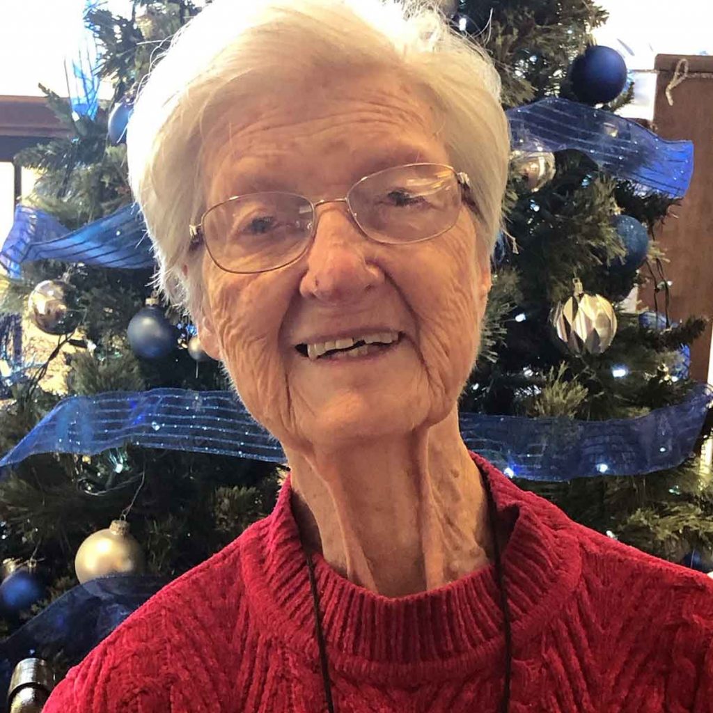 A woman with short white hair and wire-framed glasses wearing a dark red sweater and thin black lanyard smiling brightly for a headshot picture indoors in front of a blue and silver decorated Christmas tree.