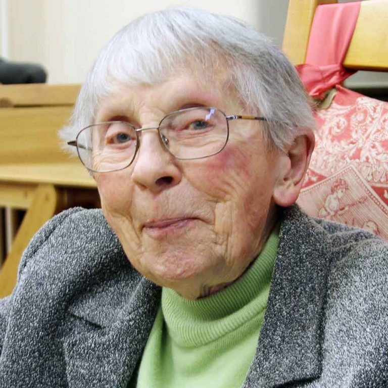 A woman with short grey hair and oval wire-framed glasses wearing a light green turtleneck and a dark grey blazer smiling softly for a headshot picture indoors.