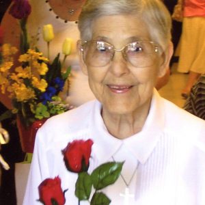 A woman with short grey hair and large clear-framed glasses wearing a white sweater with embroidered red roses smiling softly for a headshot picture at a social event.