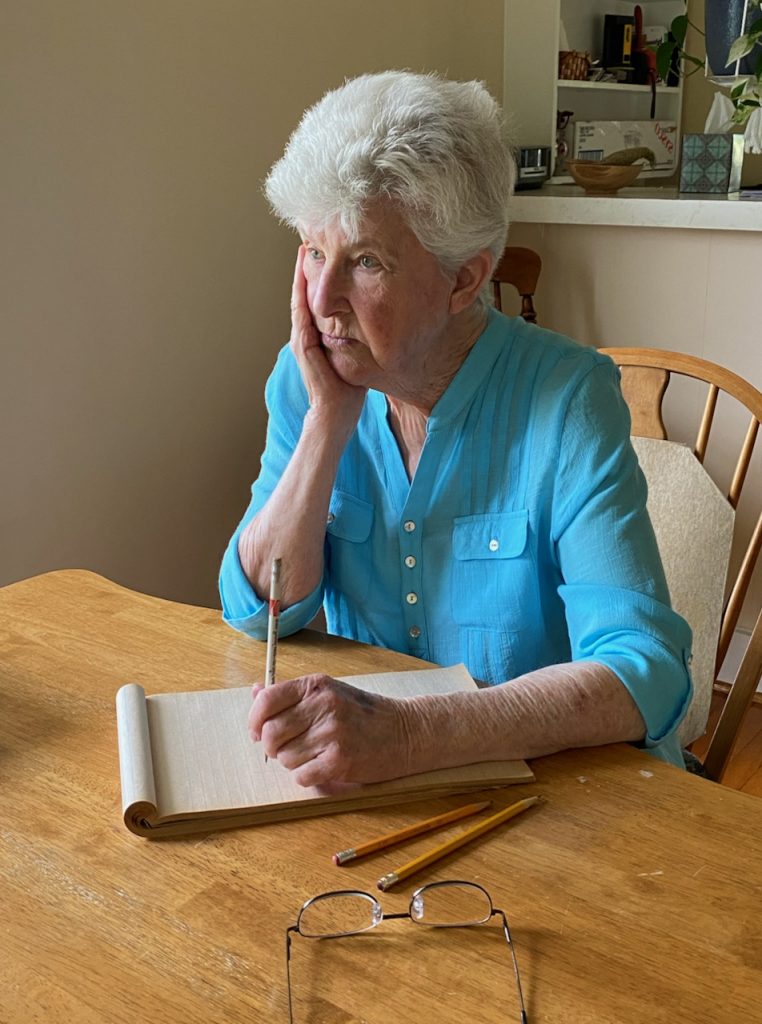 A woman sits at a table with pen and paper, contemplating what to write.