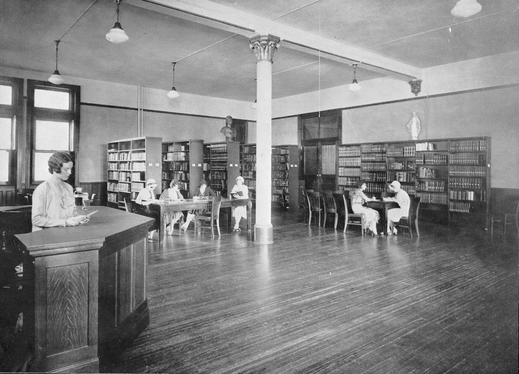 Archival photo of inside Loretto Heights College library with shelves of books and students reading at tables.