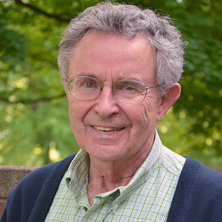 A man with short dark grey hair and round wire-framed glasses wearing a green and white plaid collared button-up shirt and a navy blue sweater smiling brightly for a headshot picture outdoors with a bright greenery background.