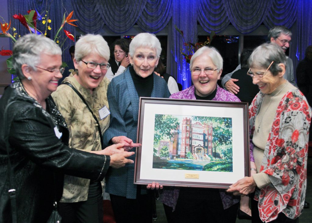 Women beaming with smiles hold a painting of Webster University building