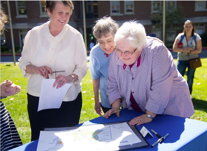 Three women stand outside looking at a drawing on a table.