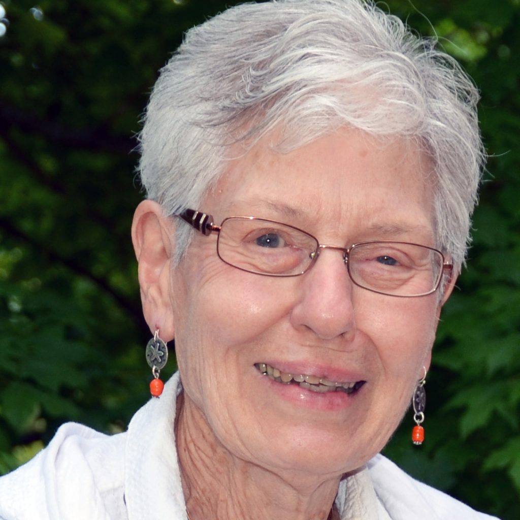 A woman with short white hair and wire-framed glasses wearing orange bead dangly earrings and a white collared blouse smiling for a headshot picture outdoors with a dark greenery background.